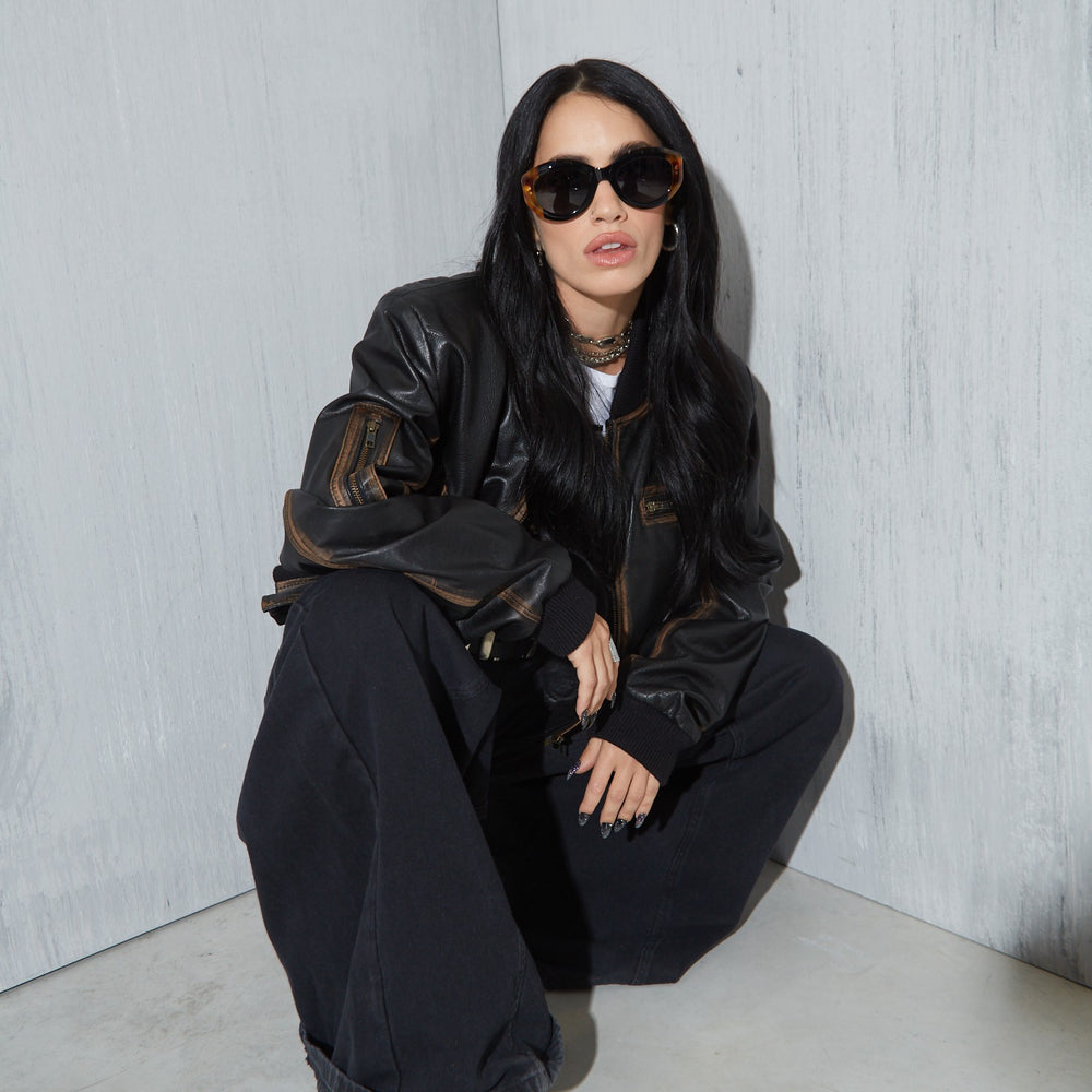Lali wearing a black leather jacket and sunglasses sitting against a light gray wall.