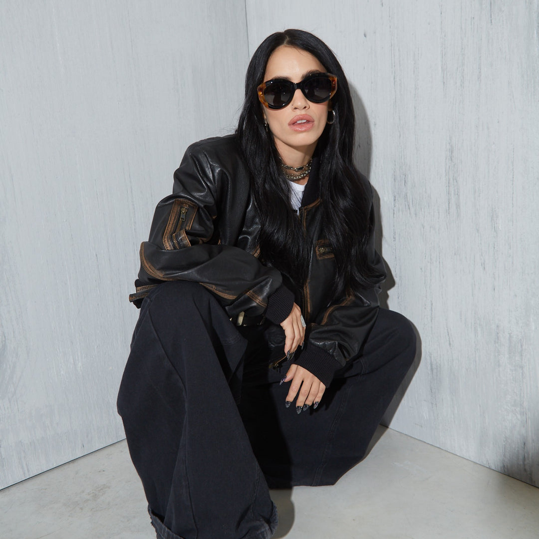 Lali wearing a black leather jacket and sunglasses sitting against a light gray wall.