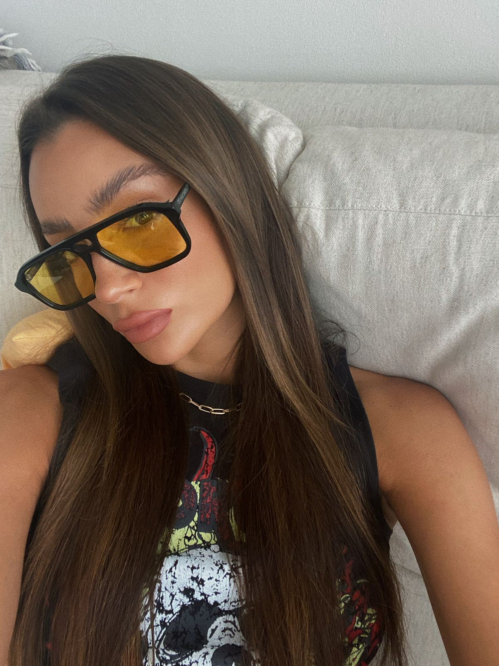 Person wearing sunglasses and a sleeveless top with a graphic design, sitting on a couch.